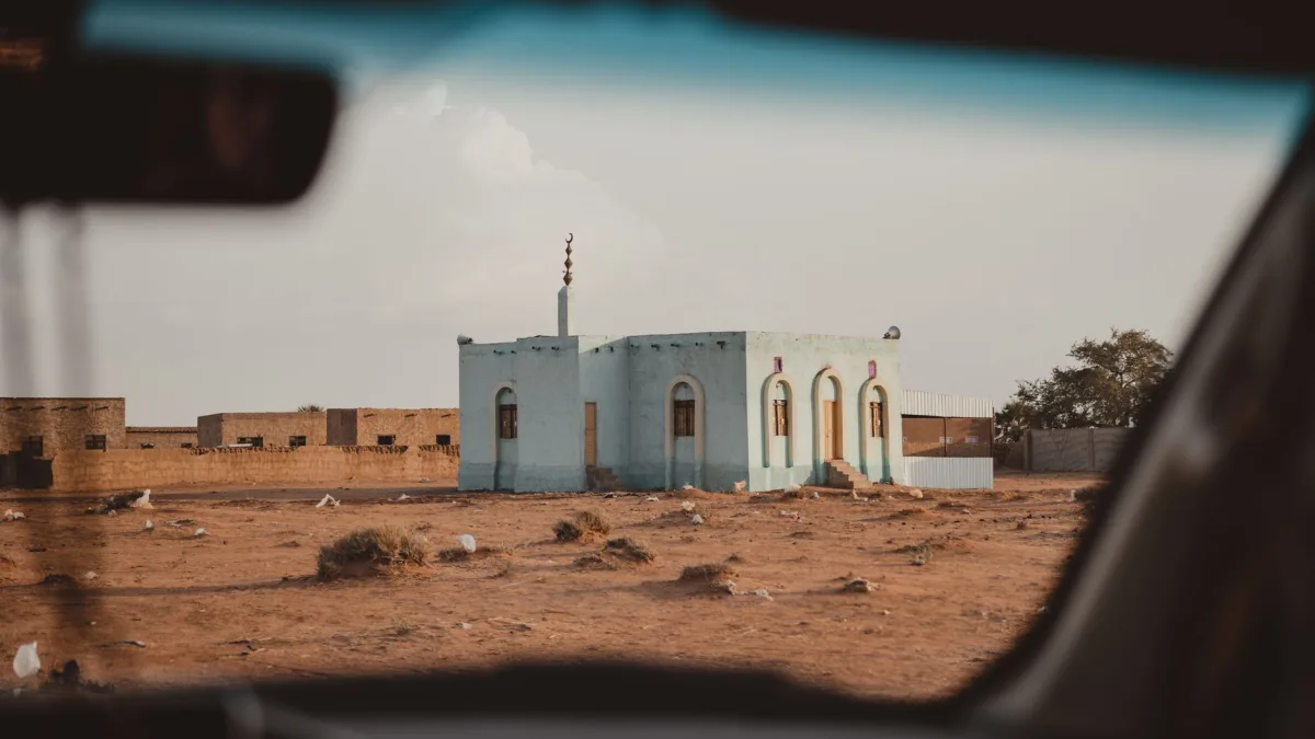 A small mosque seen through a car window in a desert landscape, under a clear sky.